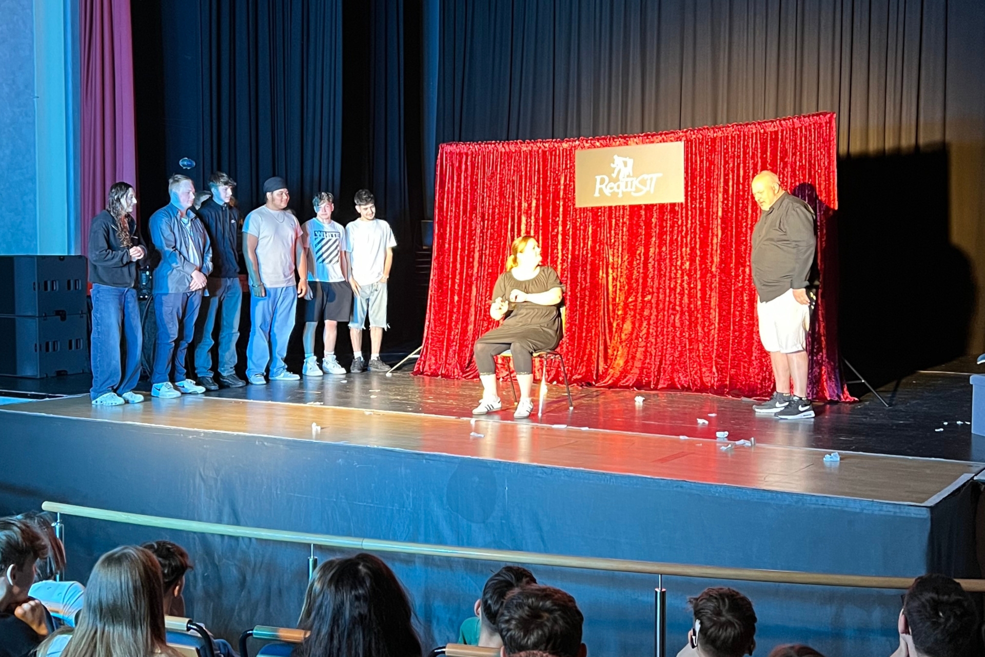 La photo montre une vue de la scène du théâtre municipal. Devant un rideau rouge, on voit deux acteurs du théâtre Requisit. A côté, six élèves regardent les acteurs.