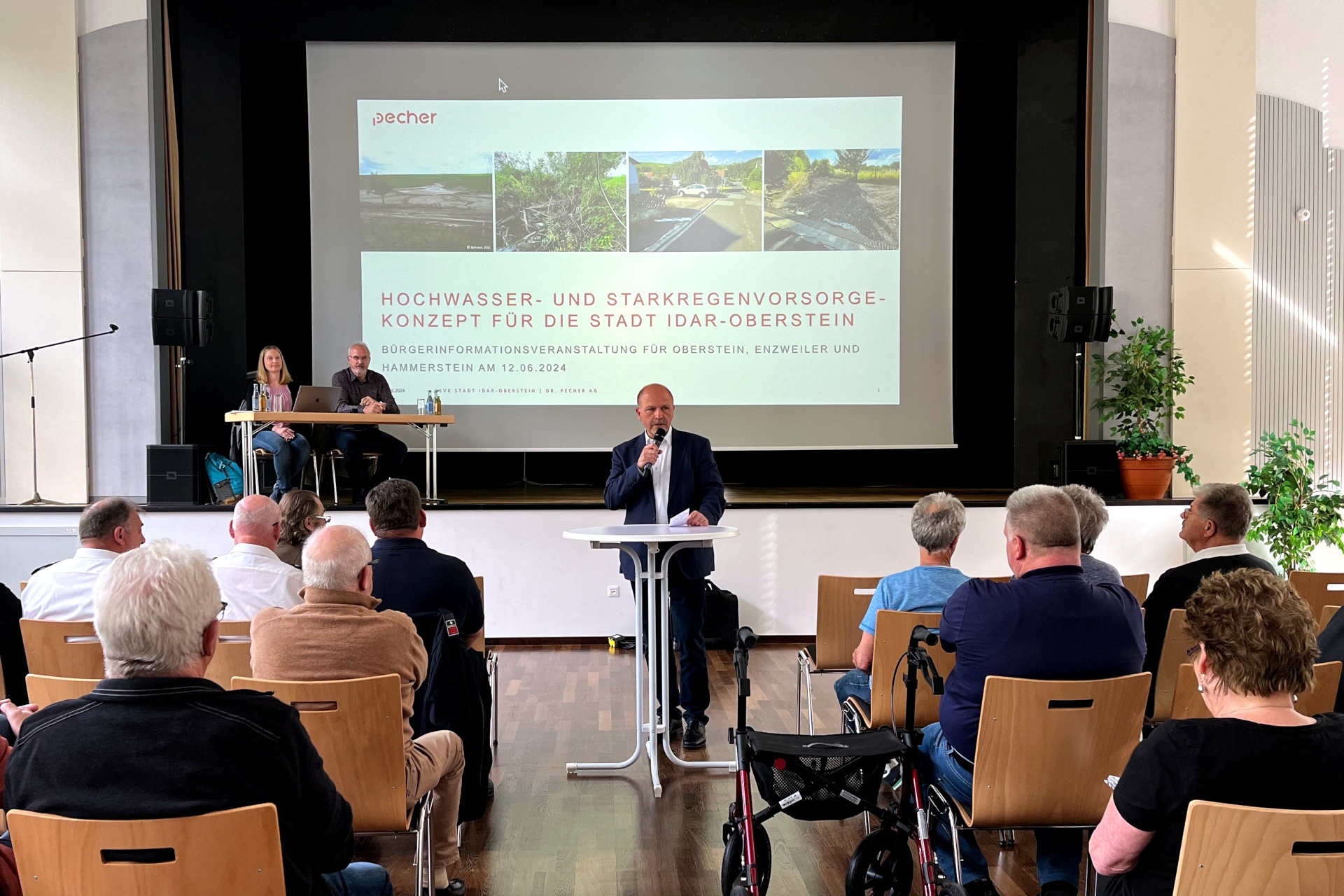 La photo montre une vue de la zone des spectateurs vers la scène. Devant la scène, le maire Marx se tient à une table haute et prononce un discours. Sur la scène, M. Webler et Mme Baron sont assis à une table à côté de l'écran sur lequel est projetée la présentation sur la DSN.