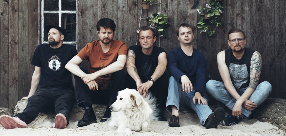 La photo montre les cinq membres du groupe. Ils sont assis côte à côte sur le sable blanc devant un hangar en bois. Un chien blanc est couché à leurs pieds.