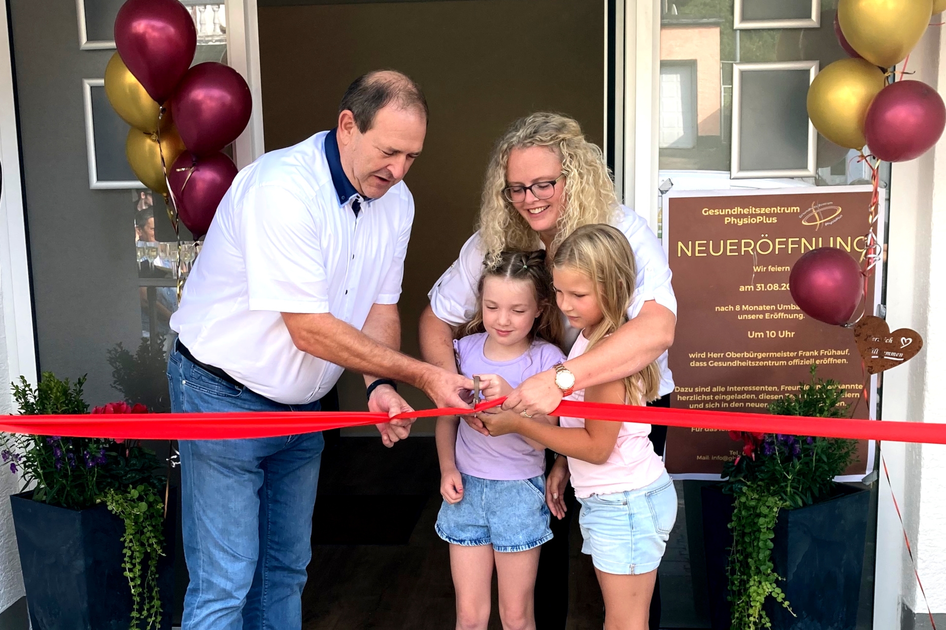 La photo montre le maire Frühauf et Franziska Schhmit ainsi que deux jeunes filles, avec l'entrée du centre de santé en arrière-plan. Les quatre personnes se tiennent derrière un ruban rouge, le maire a une paire de ciseaux à la main et s'apprête à couper le ruban.