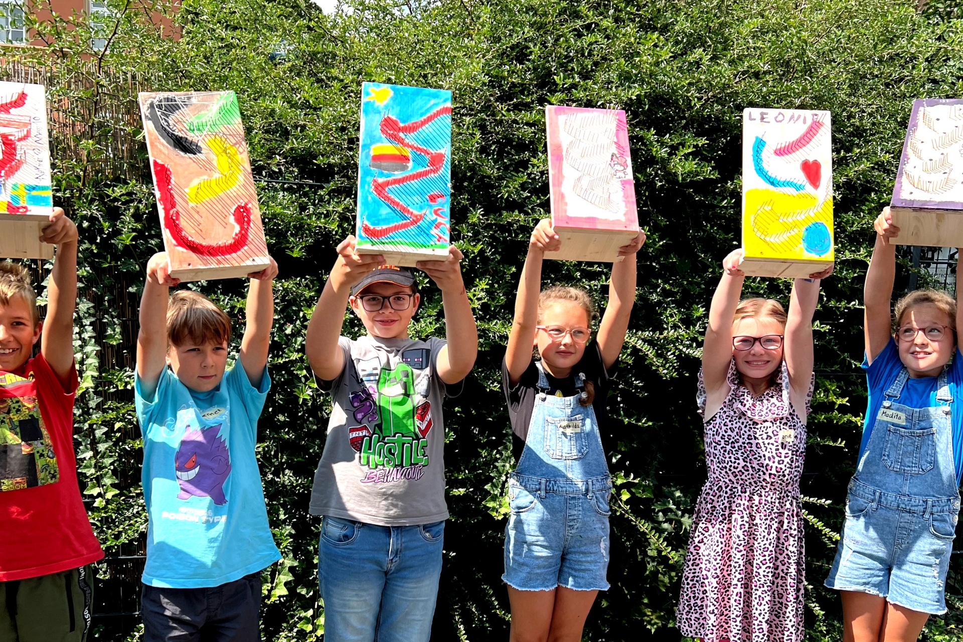 La photo montre six enfants debout côte à côte devant une haie, tenant leur circuit de billes au-dessus de leur tête.