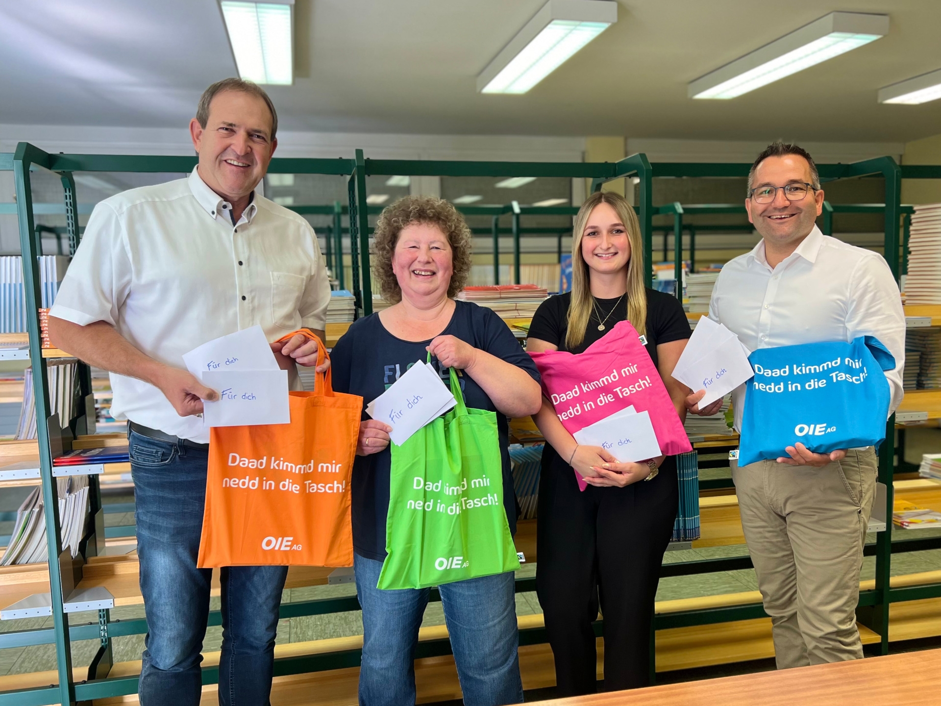 La photo montre quatre personnes debout côte à côte devant une étagère de manuels scolaires. Elles tiennent dans leurs mains les sacs en tissu multicolores de l'OIE et regardent l'appareil photo.