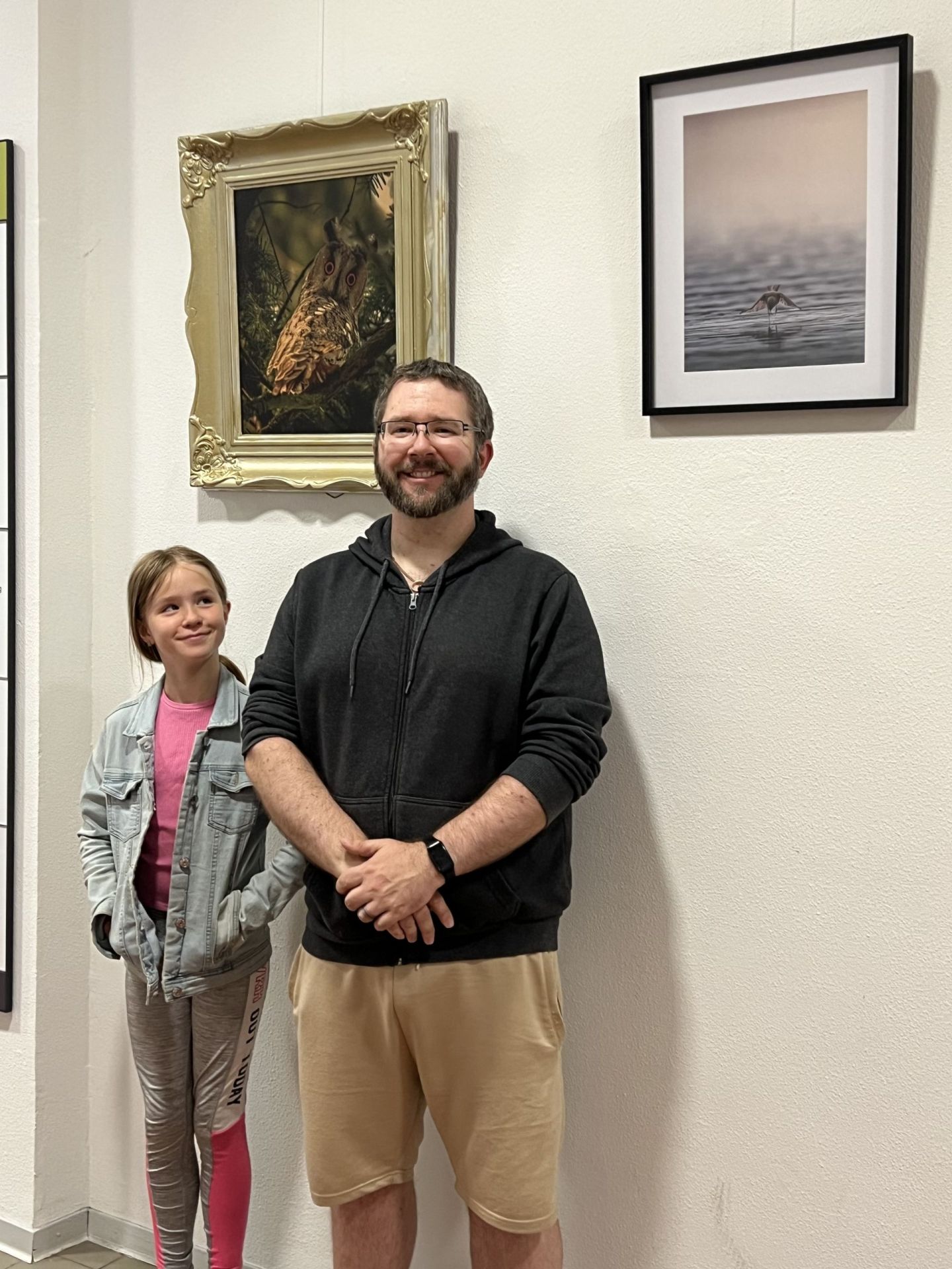 La photo montre Christopher Norman Schickling avec sa fille devant deux de ses photos.