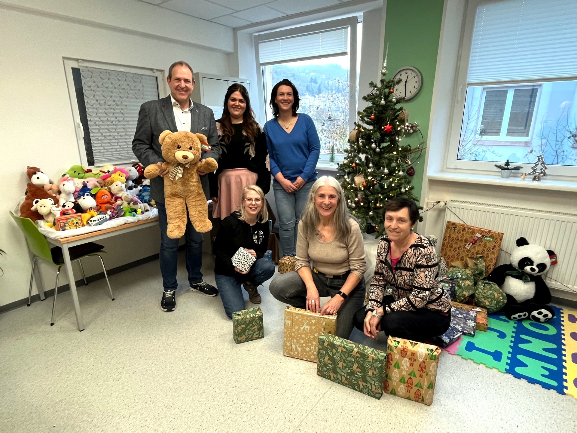 La photo montre les personnes mentionnées devant un sapin de Noël sous lequel se trouvent les cadeaux. En outre, à gauche de l'image, on peut voir la station d'adoption de doudous.