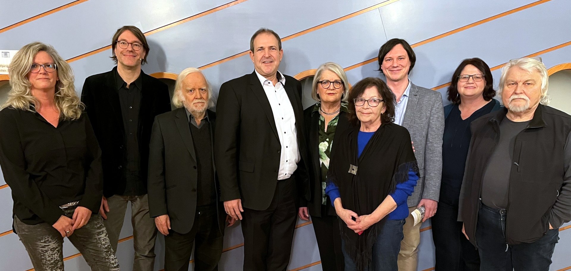 La photo montre les personnes mentionnées. Elles se tiennent côte à côte devant un mur du théâtre municipal et regardent la caméra.
