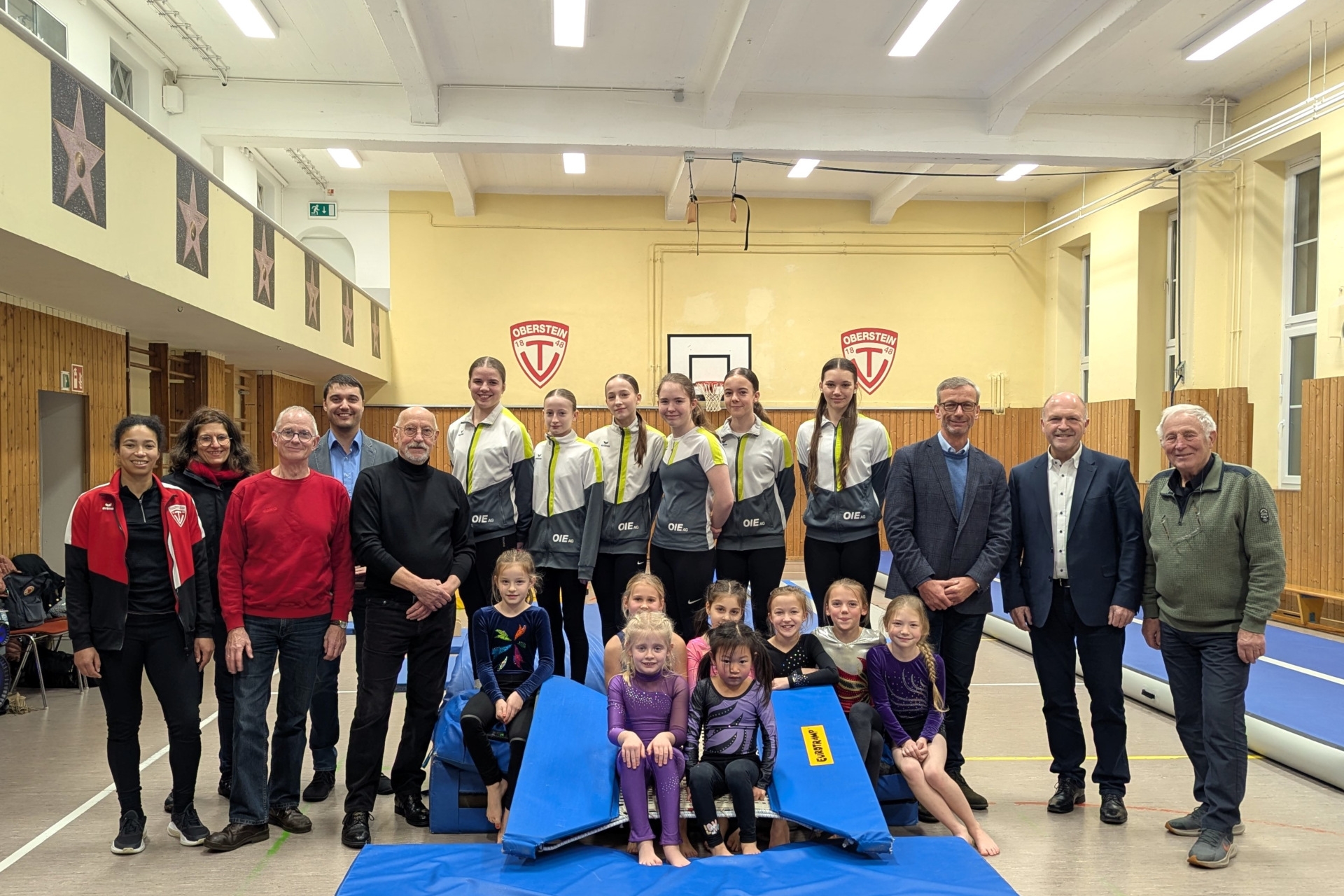 La photo montre les personnes mentionnées. Elles sont debout sur des tapis de gymnastique dans la salle de gymnastique inférieure du théâtre municipal. Au plafond, on peut voir les nouveaux éclairages LED.