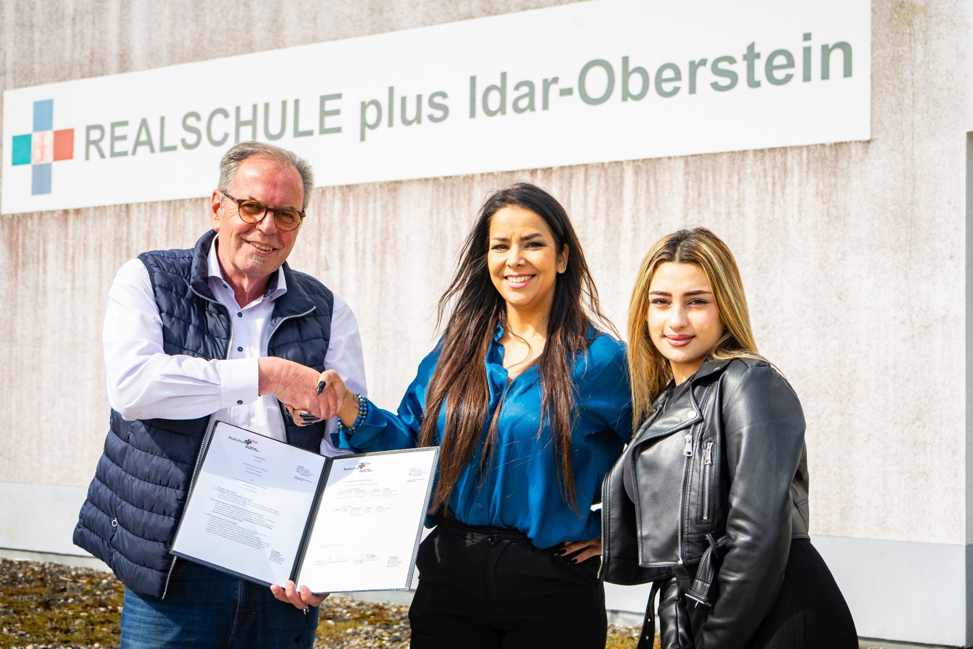 La photo montre trois personnes devant un panneau portant l'inscription Realschule plus Idar-Oberstein. Ils se serrent la main et montrent l'accord de coopération.