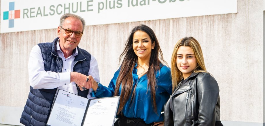La photo montre trois personnes devant un panneau portant l'inscription Realschule plus Idar-Oberstein. Ils se serrent la main et montrent l'accord de coopération.