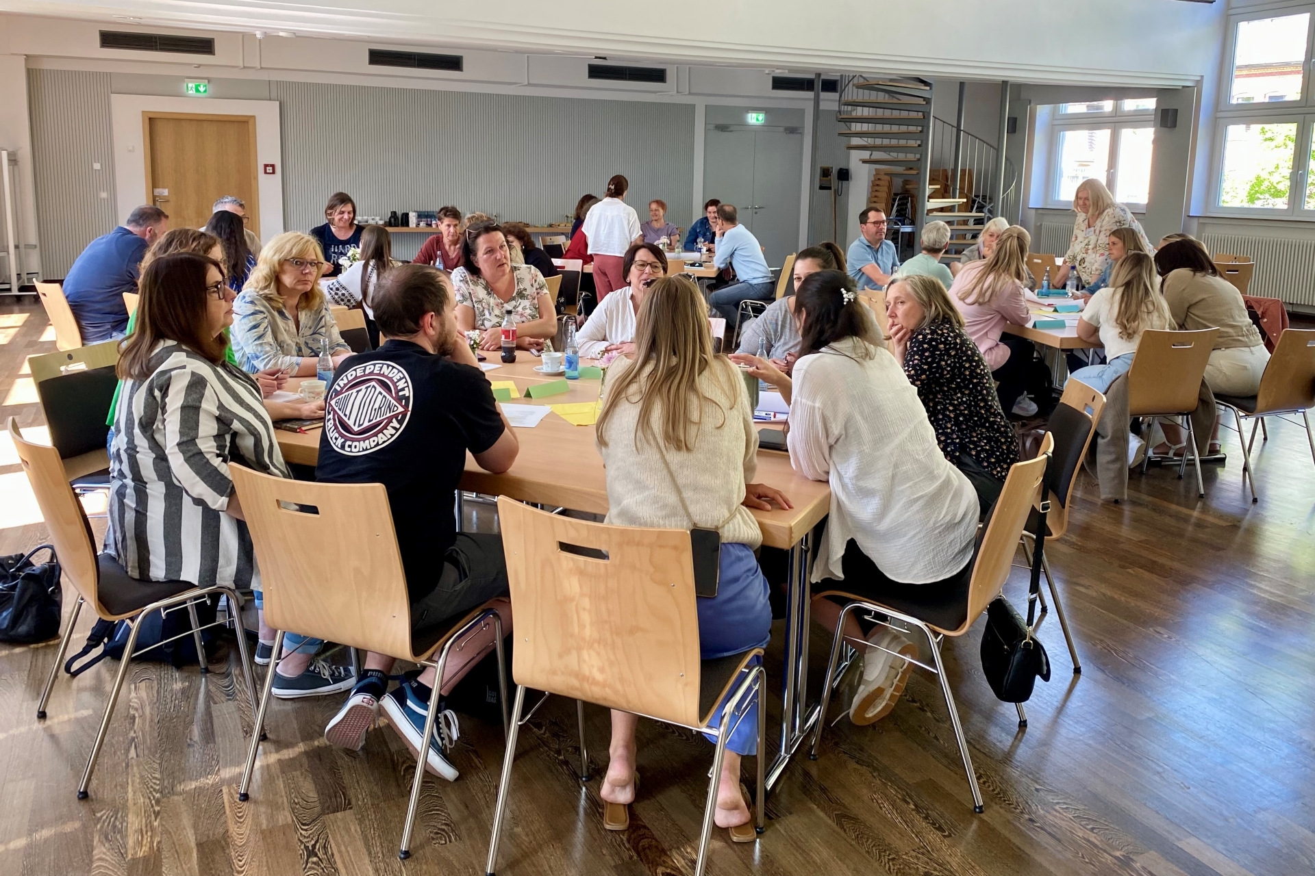 La photo montre les participants à la conférence. Ils sont assis par groupes de tables et discutent.