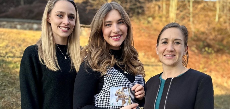 La photo montre les personnes mentionnées. Elles se tiennent côte à côte à l'extérieur et regardent la caméra. La personne du milieu tient un flyer à la main.