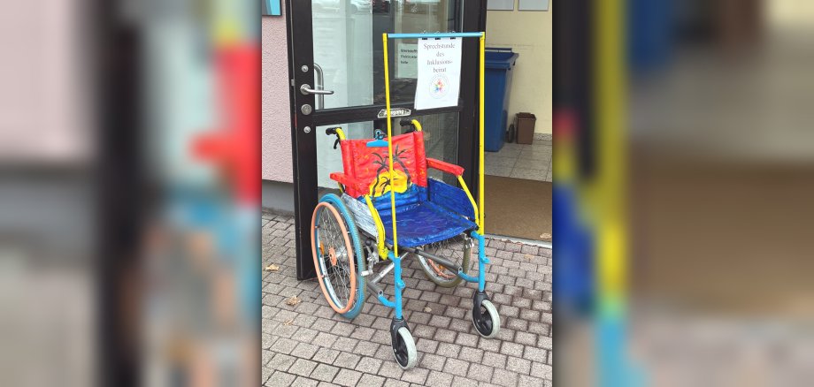 Le fauteuil roulant peint de couleurs vives attirera à l'avenir l'attention sur le travail du comité consultatif sur l'inclusion. La photo montre le fauteuil roulant coloré devant la "boîte en verre" où se tiennent les consultations du comité consultatif sur l'inclusion.