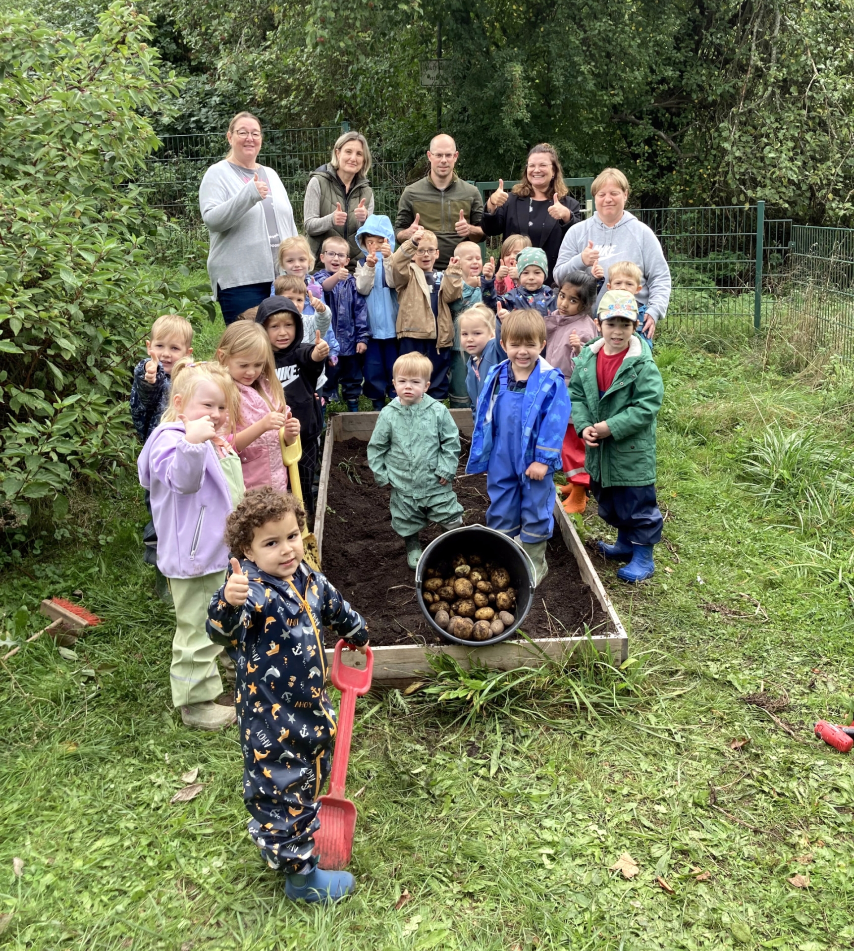 La photo montre des éducateurs, des concierges et des enfants se tenant autour du lit de plantation et présentant un seau de pommes de terre récoltées.