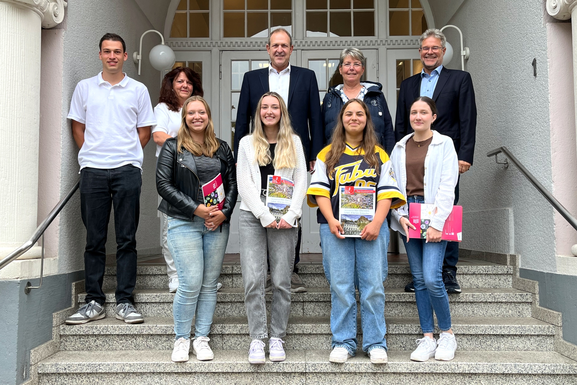 La photo montre les personnes mentionnées. Elles se tiennent en deux rangées sur les marches devant l'hôtel de ville. Les nouvelles employées se trouvent au premier rang.