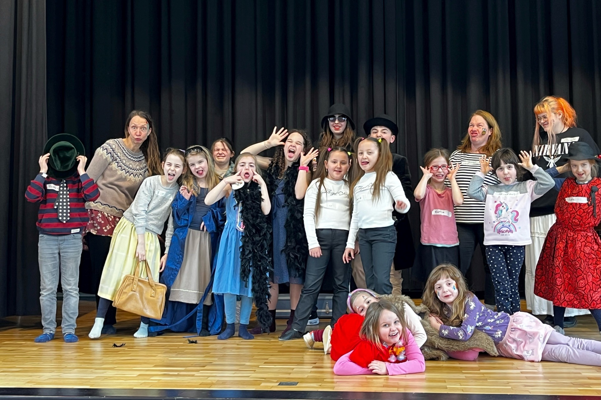 La photo montre les participants à la Journée du spectacle. Maquillés et costumés, ils se tiennent côte à côte sur la scène de l'aula Göttenbach et font des grimaces.