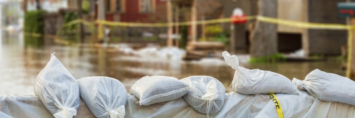 La photo montre des sacs de sable au premier plan et une rue inondée avec des maisons derrière.
