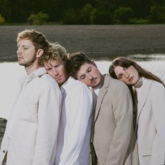 Die vier Bandmitglieder stehen hintereinander vor einem See. Sie haben jeweils den Kopf auf die Schultern der vorherigen Person gelegt.