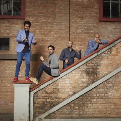 Die vier Bandmitglieder stehen und sitzen an und auf dem Geländer einer Steintreppe.
