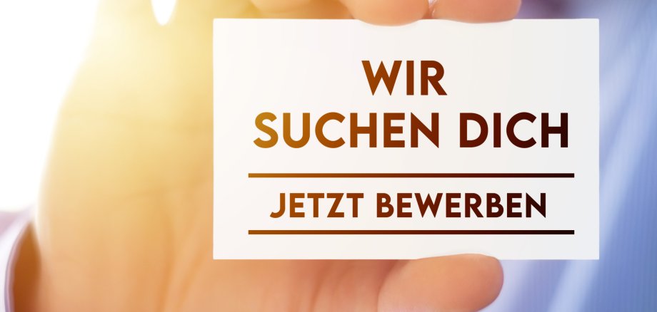 Eine Person hält eine Karte mit der Aufschrift "Wir suchen Dich - Jetzt bewerben" in die Kamera.