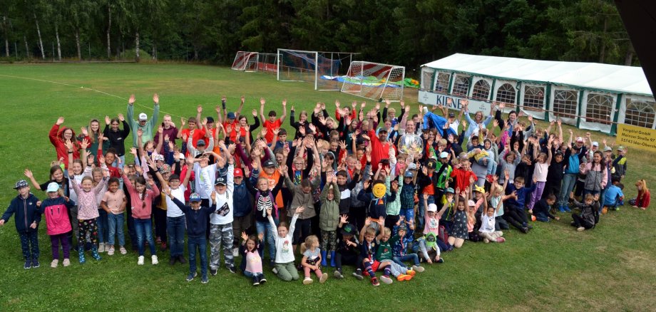 Das Foto zeigt die Kinder der Stadtranderholung mit Betreuern und Besuchern. Sie stehen alle auf dem Sportplatz und winken in die Kamera.