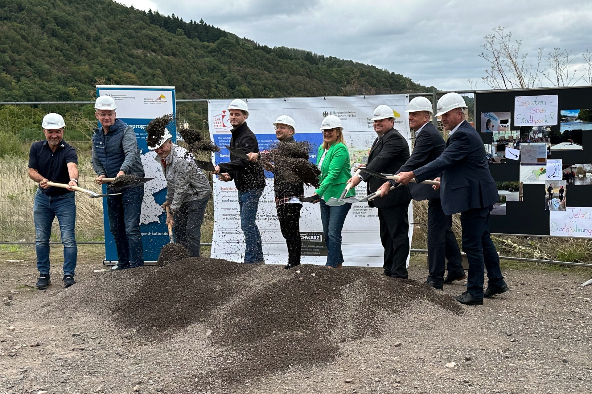 Das Foto zeigt die genannten Personen von links nach rechts. Sie stehen mit Bauhelm und Spaten an einem Sandhaufen und werfen Erde in die Luft.