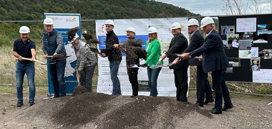 Das Foto zeigt die genannten Personen von links nach rechts. Sie stehen mit Bauhelm und Spaten an einem Sandhaufen und werfen Erde in die Luft.