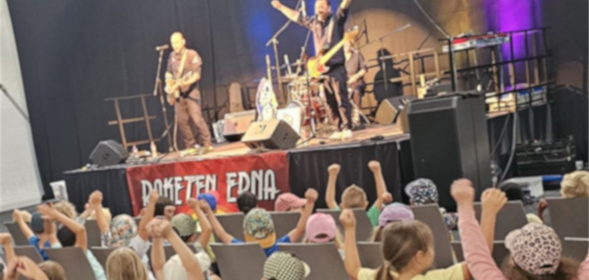 Das Foto zeigt viele Kinder die auf Stühlen vor der Bühne sitzen. Auf der Bühne spielt die Band Raketen, 