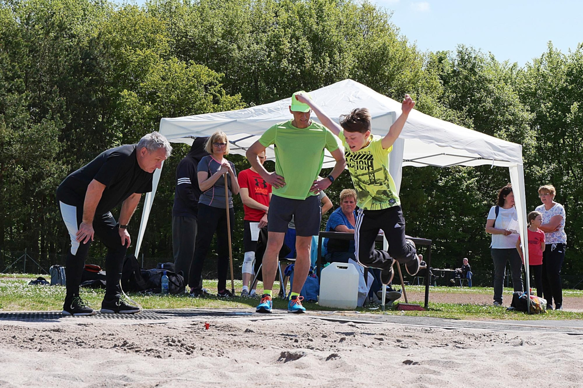 Das Foto zeigt einen Jungen beim Weitspringen. Am Rand der Sprunggrube stehen mehrere Erwachsene. Zwei Kampfrichter darauf, dass der Absprungbalken getroffen wurde, andere beobachten den Sprung.