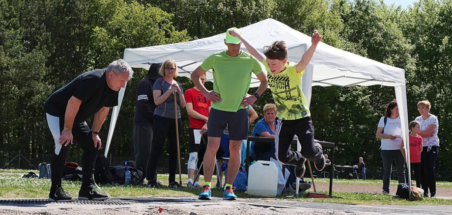Das Foto zeigt einen Jungen beim Weitspringen. Am Rand der Sprunggrube stehen mehrere Erwachsene. Zwei Kampfrichter darauf, dass der Absprungbalken getroffen wurde, andere beobachten den Sprung.