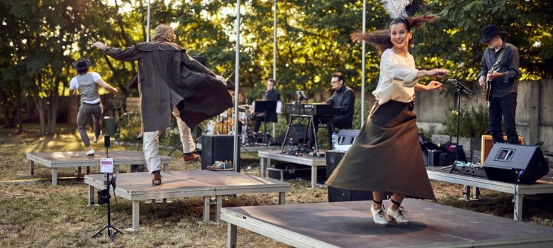 Das Foto zeigt drei Tänzer der Company, die  im Freien einzeln auf Bühnenpodesten tanzen. Dahinter sind Musiker zu sehen die ebenfalls einzeln auf Bühnenpodesten platziert sind.