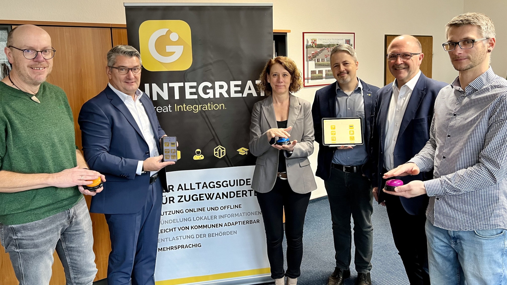 Das Foto zeigt sechs Personen, die vor einem Rollup mit der Aufschrift Integreat-App stehen. Sie halten Buzzer in den Händen, mit denen sie symbolisch die App freischalten.