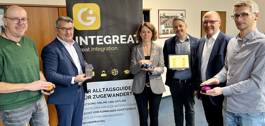 Das Foto zeigt sechs Personen, die vor einem Rollup mit der Aufschrift Integreat-App stehen. Sie halten Buzzer in den Händen, mit denen sie symbolisch die App freischalten.