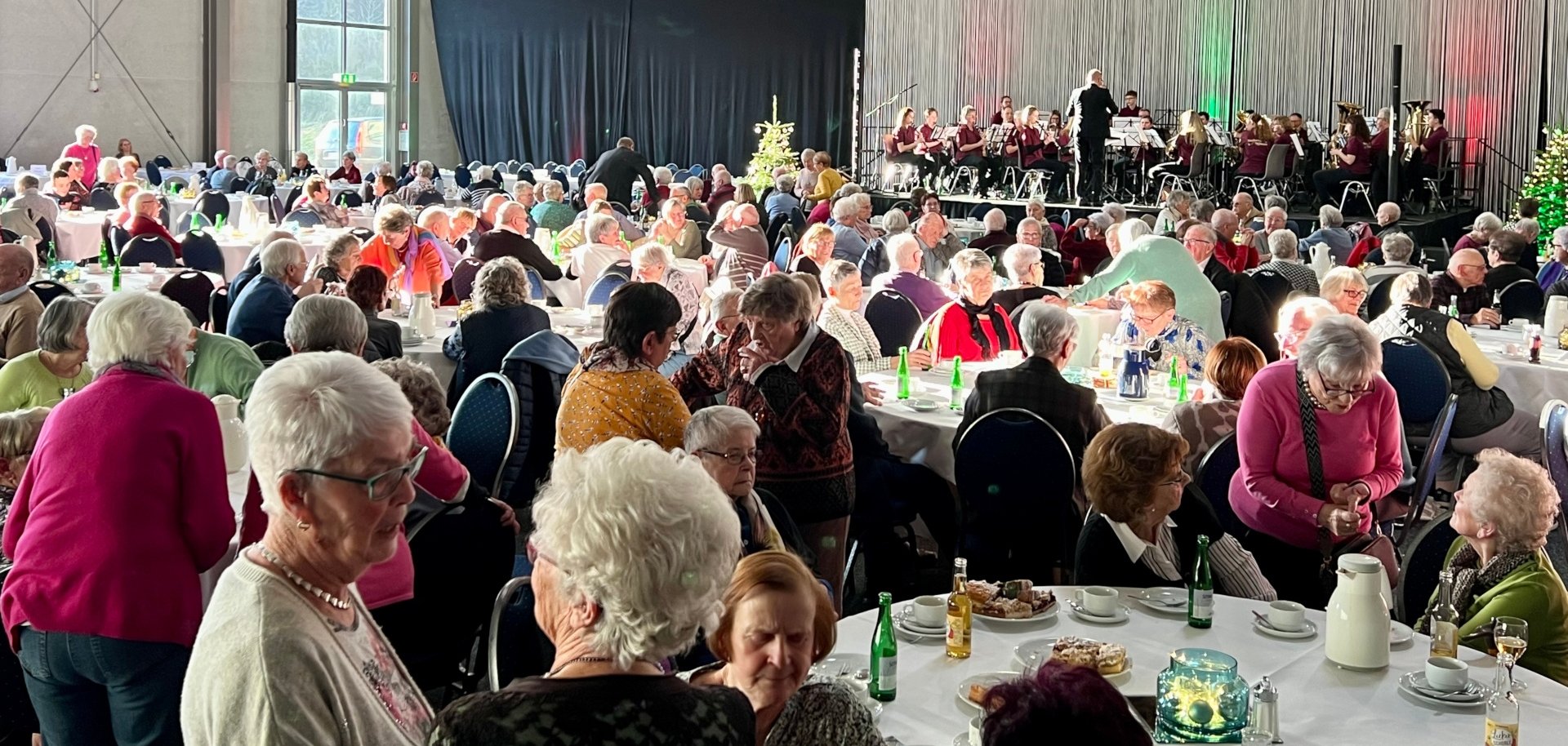 Das Foto zeigt einen Blick in die Messehalle. Zu sehen sind viele Besucher an den Tischen. Im Hintergrund ist der Musikverein auf der Bühne zu sehen.