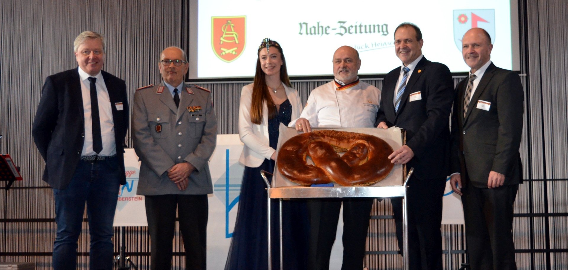 Das Foto zeigt die genannten Personen, die nebeneinander auf der Bühne stehen. Vor dem Bäckermeister steht ein Servierwagen mit der Neujahrsbrezel.