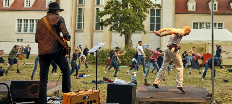 Im Vordergrund sind ein Tänzer und ein Musiker jeweils auf einem Bühnepondest zu sehen. Im Hintergrund zahlreiche Teilnehmer des Dance-Mob, die die Bewegungen des Tänzers nachahmen. 