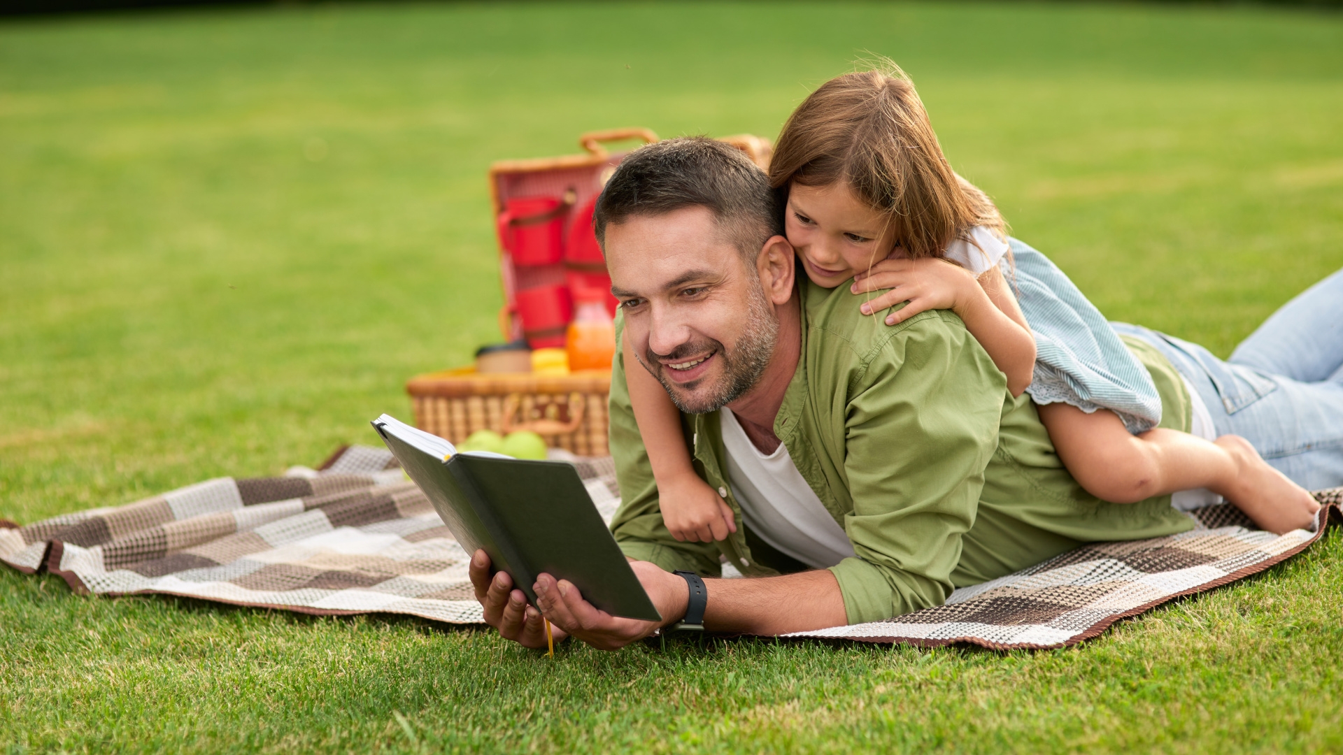 Das Foto zeigt einen Mann, der auf einer Wiese liegt und ein Buch liest. Auf seinem Rücken liegt ein Mädchen und liest mit.