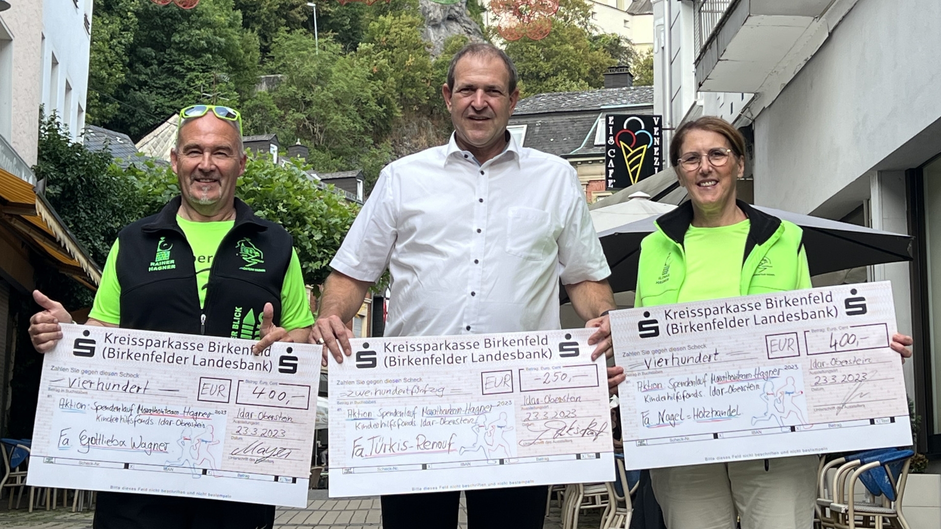 Das Foto zeigt von links nach rechts Rainer Hagner, Oberbürgermeister Frank Frühauf und Ilonka Hagner. Jeder hat einen großen Spendenscheck in den Händen, im Hintergrund ist die Felsenkirche zu sehen. 