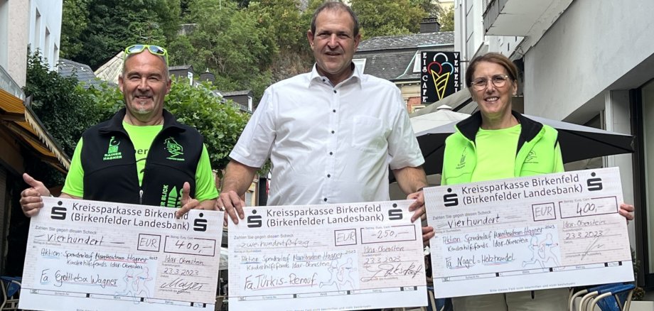 Das Foto zeigt von links nach rechts Rainer Hagner, Oberbürgermeister Frank Frühauf und Ilonka Hagner. Jeder hat einen großen Spendenscheck in den Händen, im Hintergrund ist die Felsenkirche zu sehen. 