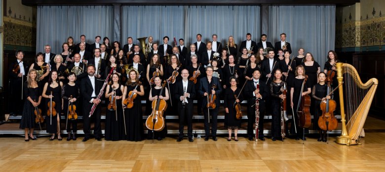 Das Foto zeigt die Mitglieder des Staatsorchester mit ihren Instrumenten in vier aufsteigenden Reihen in einem Festsaal mit Kronleuchtern.