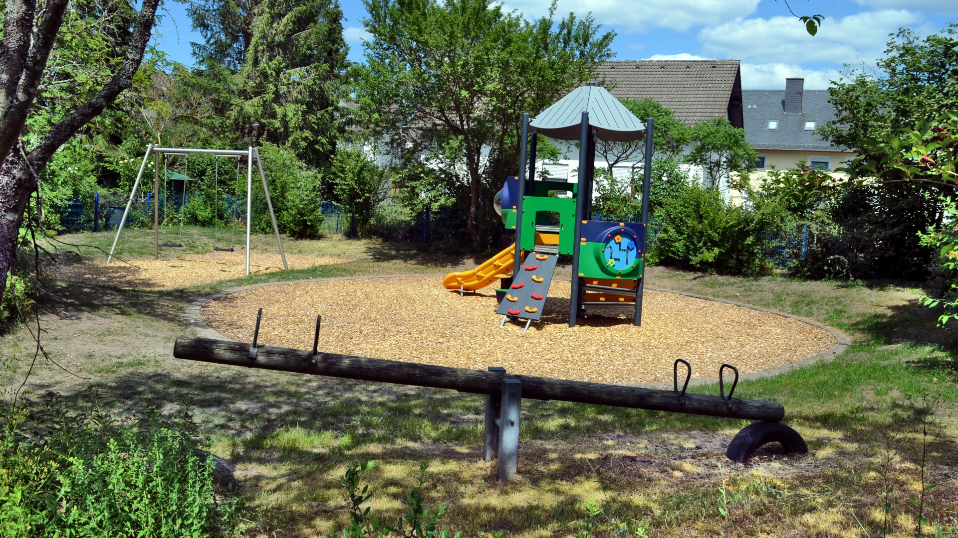 Das Foto zeigt Teile der Spielgeräte auf dem Spielplatz Wiesendellstraße. Im Vordergrund ist eine Wippe, im Hintergrund sind die neue Doppelschaukel und das neue zentrale Spielgerät zu sehen.