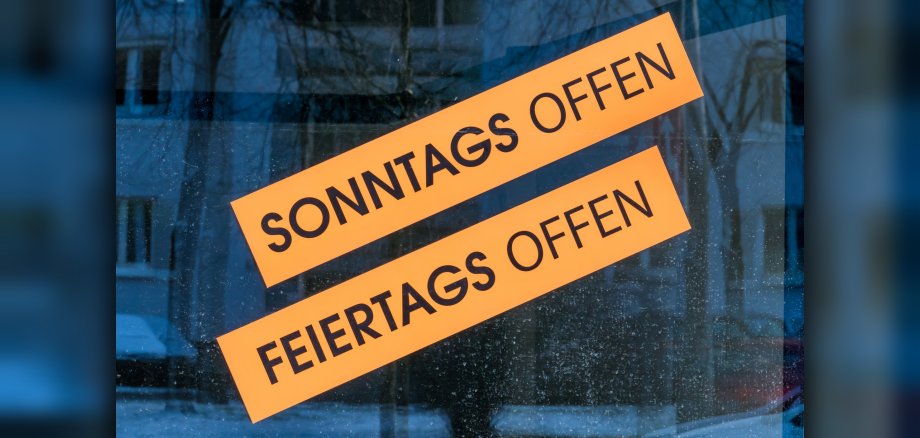 Das Foto zeigt ein Schaufenster mit den beiden Aufklebern "Sonntag geöffnet" und "Feiertag geöffnet".