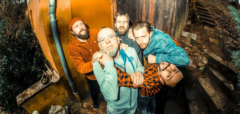 Das Foto zeigt Thomas D und die Bandmitglieder von The KBCS vor einer Hütte im Wald.