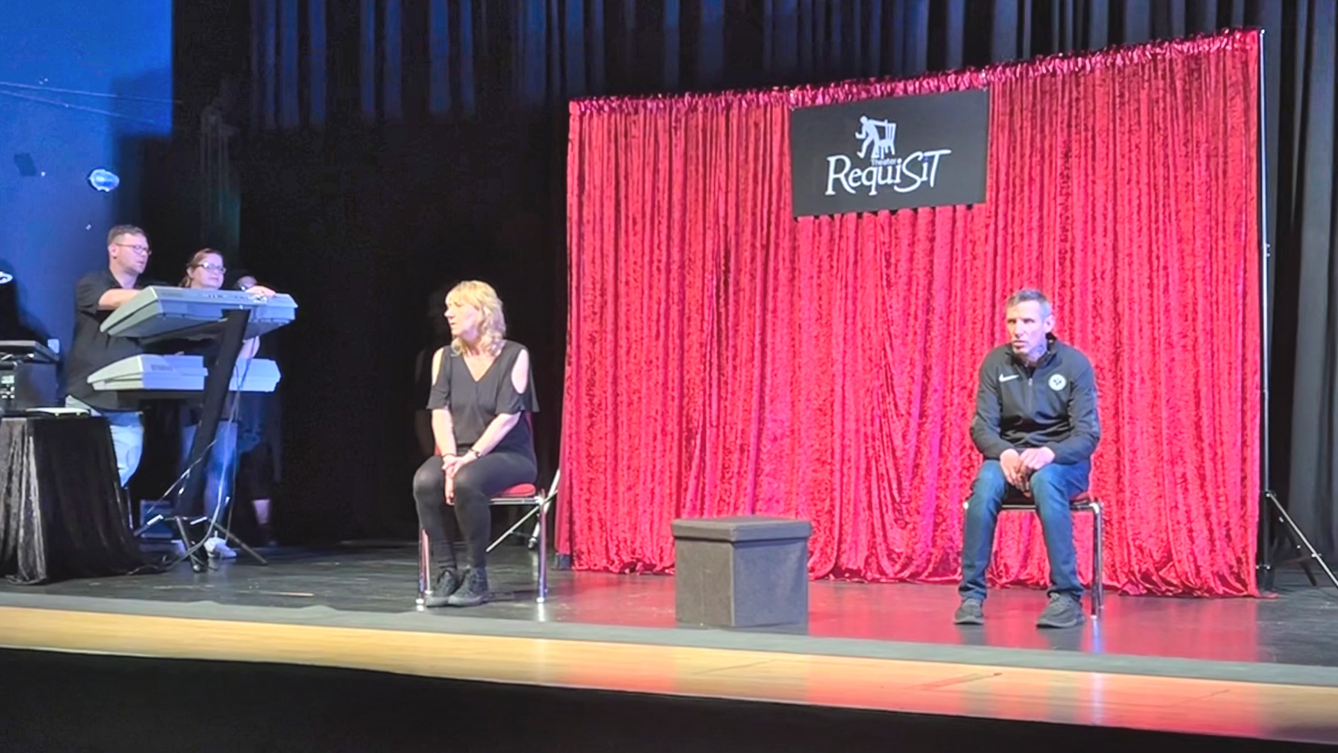 Das Foto zeigt eine Schauspielerin und einen Schauspieler des Theaters Requisit, die auf der Bühne des Stadttheaters auf Stühlen vor einem roten Vorhang sitzen. Links im Bild sind noch zwei Techniker mit ihrem Equipment zu sehen.