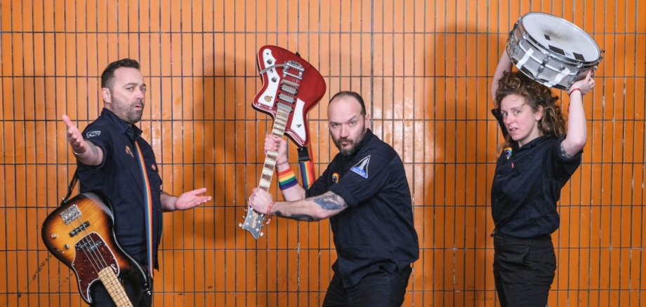 Das Foto zeigt die drei Musiker der Band Raketen Erna mit ihren Instrumenten vor einer Fliesenwand.