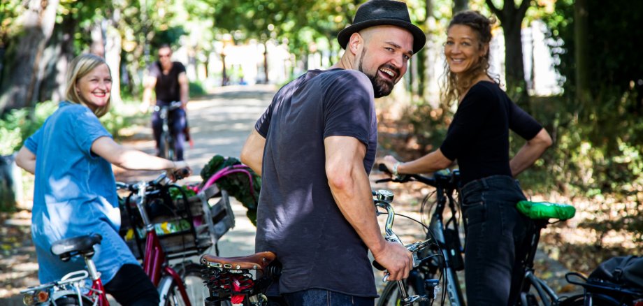 Das Foto zeigt drei Radfahrende, die mit ihren Rädern auf einem von Bäumen gesäumten Weg stehen und in die Kamera schauen.