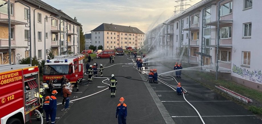 Das Foto zeigt eine Häuserzeile. Davor stehen Feuerwehrautos und die Mitglieder der Jugendfeuerwehr, die mit Strahlrohren Wasser auf die Häuser spritzen.