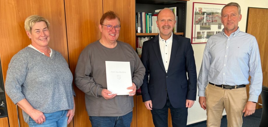 Das Foto zeigt von links Marion Müller, Rudi Bonn, Bürgermeister Friedrich Marx und Christoph Hahn (Vertreter Personalrat). Sie stehen in einer Reihe vor einer Schrankwand und blicken in die Kamera. Rudi Bonn hält die Dankurkunde in den Händen. 