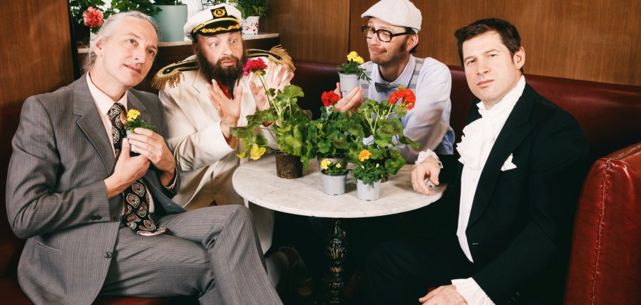 Das Foto zeigt die vier Musiker, die in Kostümen um einen runden Tisch sitzen. 
