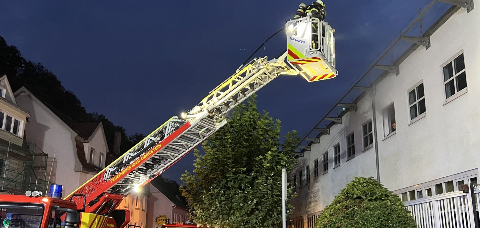 Auf dem Foto stehen zwei beleuchtete Feuerwehrfahrzeuge bei beginnender Dunkelheit vor dem OIE Gebäude. Eines davon ist die Drehleiter mit gerade zwei Feuerwehrleute in die Höhe fahren. Ein weiterer Feuerwehrmann geht auf das Gebäude zu.