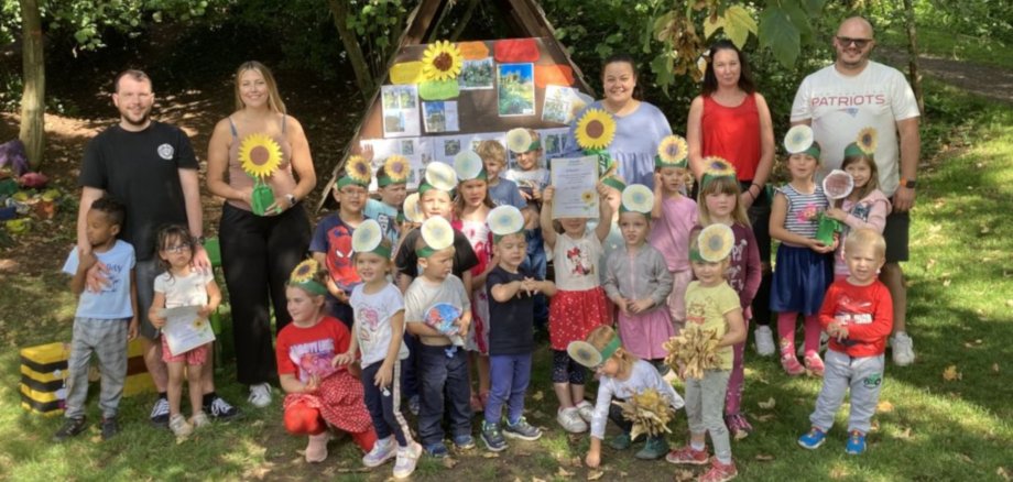 Das Foto zeigt Kinder und Erwachsene. Sie stehen mit Sonnenblumen auf einer Wiese vor einem Holzgestellt, im Hintergrund sind Bäume zu sehen.