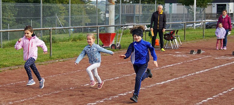 Das Foto zeigt drei Kinder, die auf der Laufbahn laufen. Im Hintergrund sind die Zeitnehmer und zwei Zuschauer zu sehen.